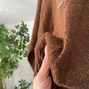 May include: A close-up of a brown knit sweater. The sweater has a textured appearance and a ribbed cuff. A small white emblem is visible on the upper left side of the sweater. The sweater is made of a soft, warm material.