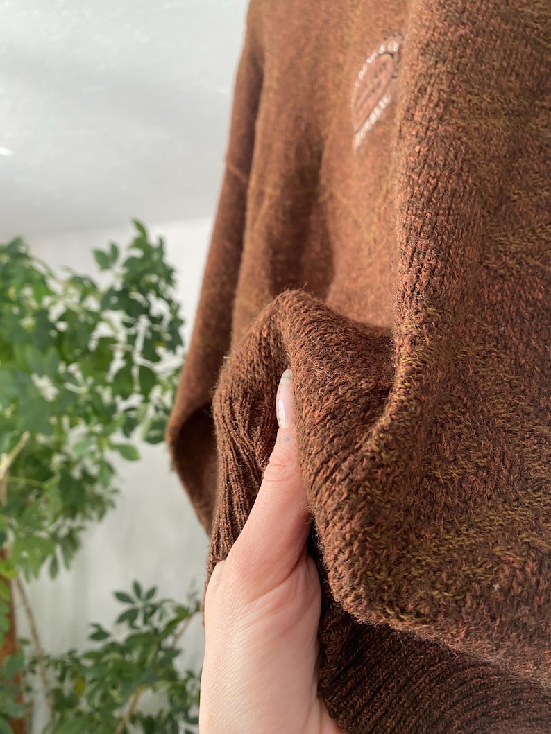 May include: A close-up of a brown knit sweater. The sweater has a textured appearance and a ribbed cuff. A small white emblem is visible on the upper left side of the sweater. The sweater is made of a soft, warm material.