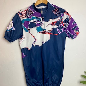 May include: A navy blue cycling jersey with a white, purple, red, and teal abstract design. The jersey has a half-zip closure and short sleeves.