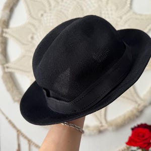 May include: A black fedora hat with a mesh-like texture and a black band around the crown. The hat has a classic, structured shape with a slightly curved brim. The hat is a stylish accessory.