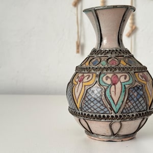 Puede incluir: Un jarrón decorativo con cuerpo rosa claro y borde de metal plateado. El jarrón presenta coloridos patrones geométricos en amarillo, azul y rojo. El jarrón mide aproximadamente 15 cm de alto.
