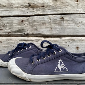May include: A pair of low-top navy blue canvas sneakers featuring dark blue laces, white rubber toe caps, and white rubber soles. A white embroidered logo of a rooster inside a triangle is visible on the side of the shoe near the heel area.