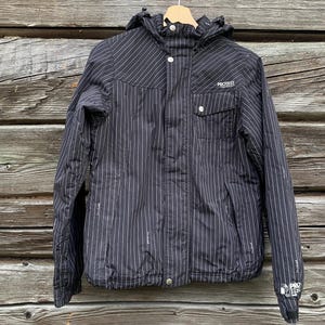 May include: Black and white striped hooded jacket with the word "PROTEST" above the chest pocket. The jacket has a zipper closure, two front pockets, and a small pocket on the left sleeve. The jacket is hanging on a wooden hanger.