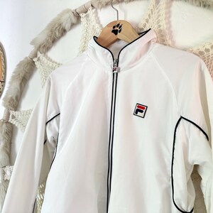 May include: White athletic jacket with black trim and a red, white, and blue logo on the chest. The jacket has a full zipper and a high collar. A vintage tennis racket hangs in the background.