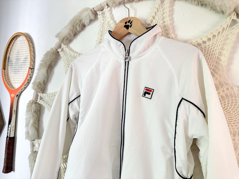 May include: White athletic jacket with black trim and a red, white, and blue logo on the chest. The jacket has a full zipper and a high collar. A vintage tennis racket hangs in the background.