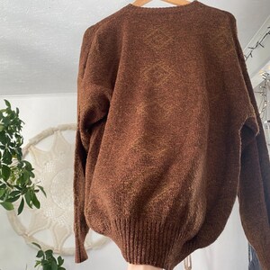 May include: A brown knit sweater with a ribbed hem and cuffs. The sweater has a V-neck and long sleeves. The fabric has a textured appearance with subtle variations in color. The sweater is hanging on a wooden hanger.