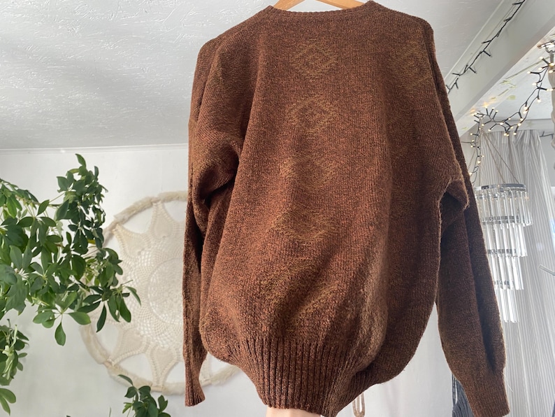 May include: A brown knit sweater with a ribbed hem and cuffs. The sweater has a V-neck and long sleeves. The fabric has a textured appearance with subtle variations in color. The sweater is hanging on a wooden hanger.