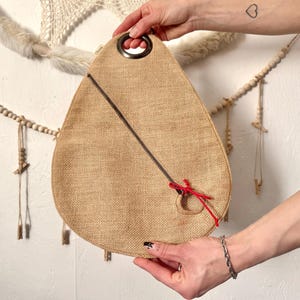 May include: A tan burlap bag with a black zipper and a red accent. The bag has a dark metal ring handle and a circular opening with a red tie. The bag is held up by two hands.