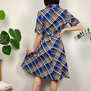 May include: A blue, brown, and white plaid dress with a short sleeve and a fitted bodice. The dress has a flared skirt and a belt.