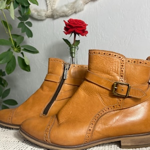 K&ouml;nnte beinhalten: Ein Paar hellbraune Leder-Ankle-Boots mit dekorativen N&auml;hten und Schnallenverschluss. Die Stiefel haben einen seitlichen Rei&szlig;verschluss und einen niedrigen Absatz. Eine rote Rose in einer kleinen Vase im Hintergrund.
