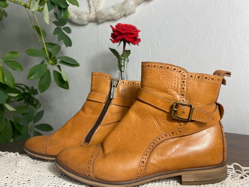 K&ouml;nnte beinhalten: Ein Paar hellbraune Leder-Ankle-Boots mit dekorativen N&auml;hten und Schnallenverschluss. Die Stiefel haben einen seitlichen Rei&szlig;verschluss und einen niedrigen Absatz. Eine rote Rose in einer kleinen Vase im Hintergrund.