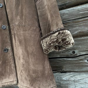 May include: A brown suede coat with a faux fur trim on the cuffs. The coat has a button-up front and a classic design. The image shows a close-up of the coat's details, including the texture of the suede and the fur.