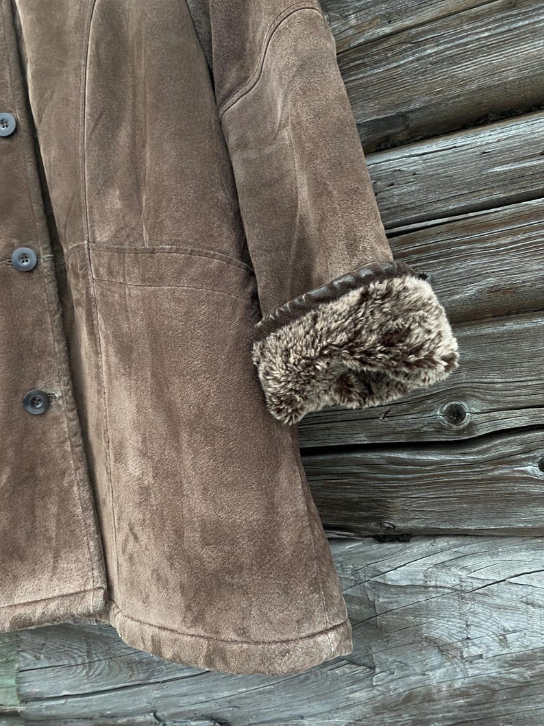 May include: A brown suede coat with a faux fur trim on the cuffs. The coat has a button-up front and a classic design. The image shows a close-up of the coat's details, including the texture of the suede and the fur.
