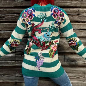 Può includere: Un maglione in maglia a maniche lunghe con righe orizzontali color verde acqua e crema. Il retro è decorato con grandi applicazioni floreali colorate di paillettes e un motivo centrale di drago di paillettes nei toni del rosso, blu e verde.