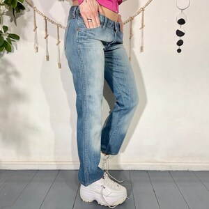 May include: Light blue denim jeans with a faded wash. The jeans have a classic five-pocket design and a straight leg fit. The model is wearing white platform sneakers. The jeans are a medium wash with some fading.