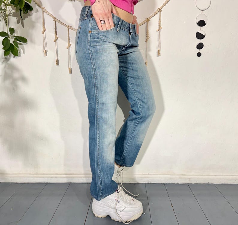 May include: Light blue denim jeans with a faded wash. The jeans have a classic five-pocket design and a straight leg fit. The model is wearing white platform sneakers. The jeans are a medium wash with some fading.