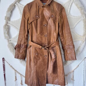 Può includere: Un trench coat in pelle marrone con cintura in vita e chiusura con bottoni. Il cappotto presenta un collo, maniche lunghe con polsini regolabili e un classico design da trench coat. Il cappotto è appeso a una gruccia di legno.
