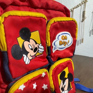 May include: Red backpack with yellow zippers and multiple pockets featuring Mickey Mouse graphics. One pocket has the text "Oh Boy!" in a speech bubble. The backpack has blue straps and is a Disney-themed item.