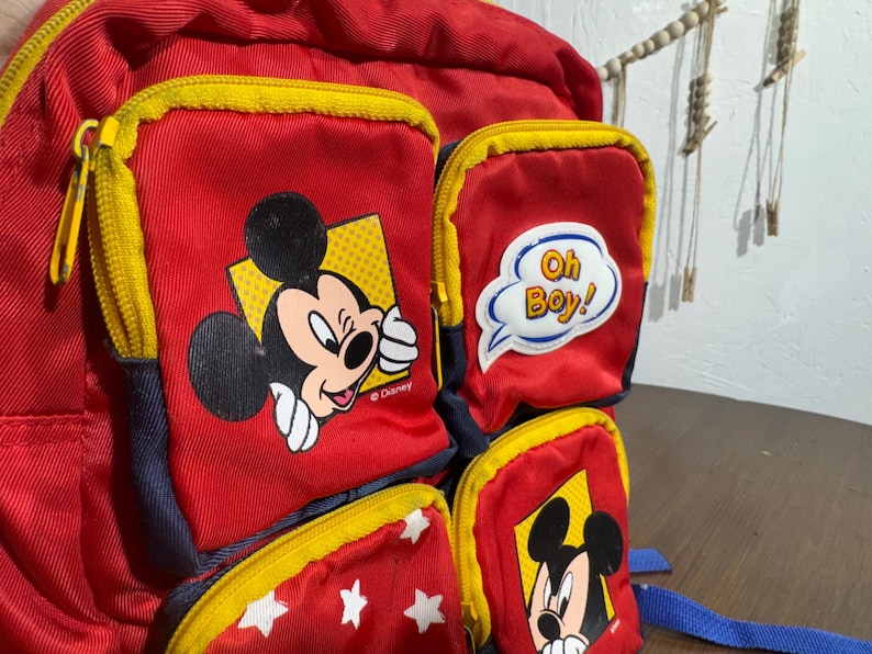 May include: Red backpack with yellow zippers and multiple pockets featuring Mickey Mouse graphics. One pocket has the text "Oh Boy!" in a speech bubble. The backpack has blue straps and is a Disney-themed item.