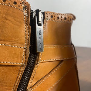 K&ouml;nnte beinhalten: Nahaufnahme eines hellbraunen Lederstiefels mit Rei&szlig;verschluss und dem Markennamen "Marc O'Polo" auf dem Rei&szlig;verschlussanh&auml;nger. Der Stiefel hat dekorative N&auml;hte und einen Riemen. Das Leder scheint leicht abgenutzt zu sein.