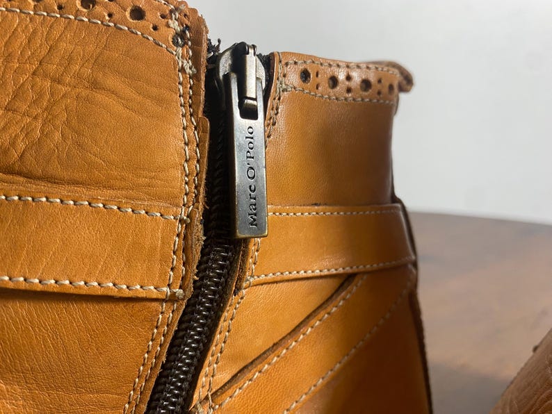 K&ouml;nnte beinhalten: Nahaufnahme eines hellbraunen Lederstiefels mit Rei&szlig;verschluss und dem Markennamen "Marc O'Polo" auf dem Rei&szlig;verschlussanh&auml;nger. Der Stiefel hat dekorative N&auml;hte und einen Riemen. Das Leder scheint leicht abgenutzt zu sein.