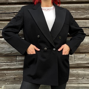 Puede incluir: Un blazer negro de doble botonadura con solapas de satén, cuatro botones frontales y dos bolsillos inferiores. El blazer se lleva sobre una camisa blanca con texto bordado en rojo cerca del cuello y presenta mangas largas.