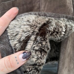 May include: Close-up of a brown suede jacket with a plush, faux fur lining. The jacket's interior features a soft, textured fur in shades of gray and brown, providing warmth and comfort. The outer suede has a matte finish.