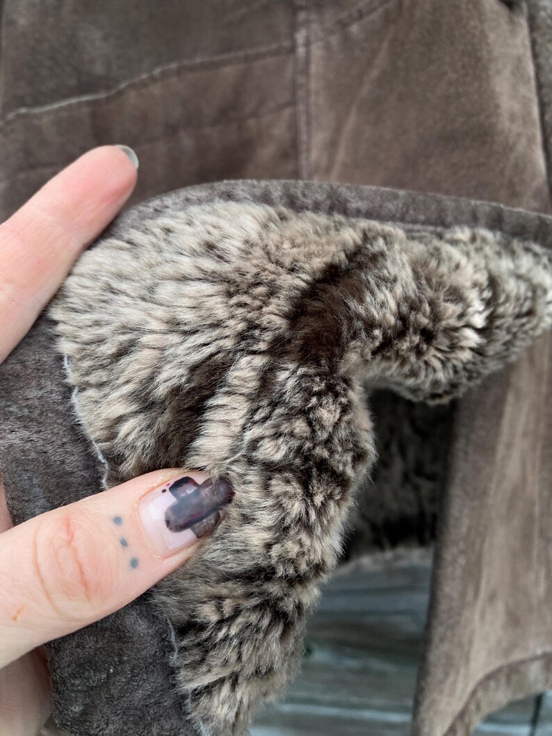 May include: Close-up of a brown suede jacket with a plush, faux fur lining. The jacket's interior features a soft, textured fur in shades of gray and brown, providing warmth and comfort. The outer suede has a matte finish.