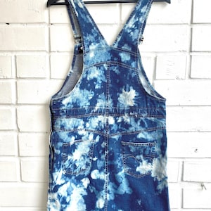 Puede incluir: Un vestido de peto vaquero azul con estampado tie-dye. El vestido tiene tirantes ajustables y dos bolsillos en la espalda.