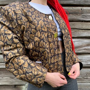 May include: A brown and gold leaf patterned jacket with gold buttons. The jacket has a round neckline and long sleeves.