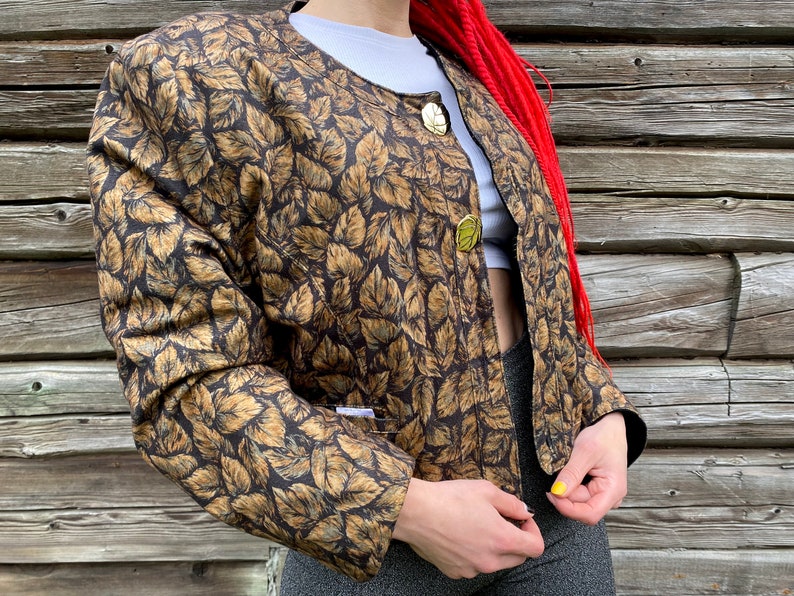 May include: A brown and gold leaf patterned jacket with gold buttons. The jacket has a round neckline and long sleeves.