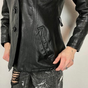 May include: A black leather jacket with a double-breasted closure and two buttons. The jacket has a pocket on the left side and a fitted silhouette.
