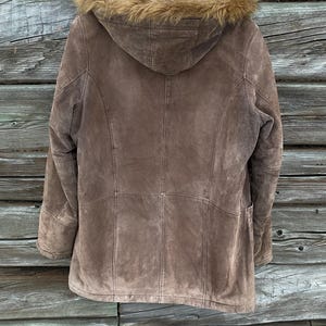 May include: A brown suede jacket with a fur-lined hood, hanging against a weathered wooden background. The jacket features a tailored design with visible stitching and a warm, neutral color palette. The fur trim adds a touch of luxury to this outerwear piece.