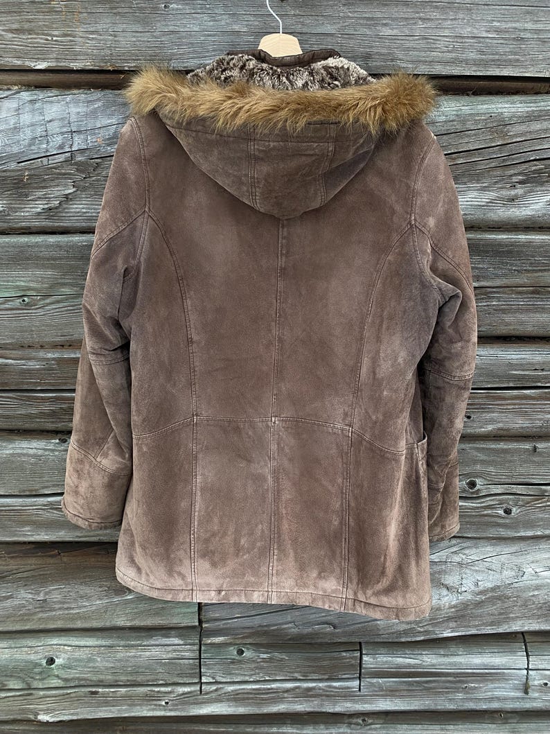 May include: A brown suede jacket with a fur-lined hood, hanging against a weathered wooden background. The jacket features a tailored design with visible stitching and a warm, neutral color palette. The fur trim adds a touch of luxury to this outerwear piece.