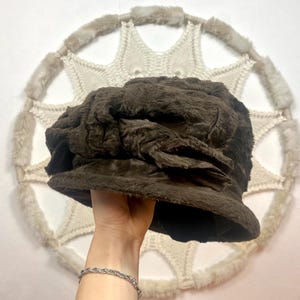 Può includere: Un cappello marrone scuro e strutturato con una corona arrotondata e un fiocco decorativo. Il cappello è mostrato davanti a un arazzo da parete circolare bianco tessuto con finiture in pelliccia.