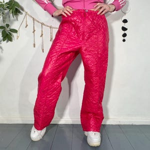 Może przedstawiać: Jasnoróżowe pikowane spodnie śniegowe. Spodnie mają wysoki stan i wzór w romby. Spodnie są pokazane z białymi trampkami i różową kurtką. Spodnie są wykonane z błyszczącego materiału.