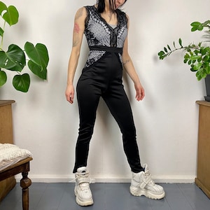 Puede incluir: Un mono negro con una superposición de encaje y una silueta ajustada. El mono tiene un escote en V y una pierna ancha. La modelo lleva zapatillas blancas de plataforma.