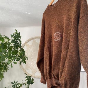May include: A brown sweater with a crew neck and long sleeves hangs from a wooden hanger. The sweater has a textured appearance and features a circular emblem on the chest with the text "ENGLISH STONE". The sweater is a warm, neutral color.