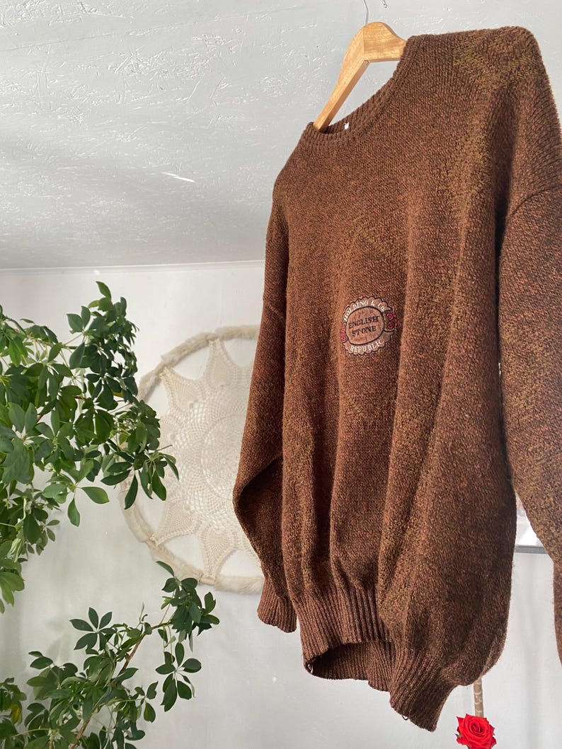 May include: A brown sweater with a crew neck and long sleeves hangs from a wooden hanger. The sweater has a textured appearance and features a circular emblem on the chest with the text "ENGLISH STONE". The sweater is a warm, neutral color.