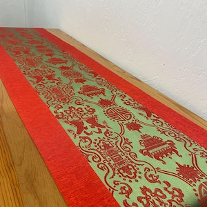 May include: A green and red table runner with a traditional Chinese design. The runner has a red border and a green center with intricate patterns.