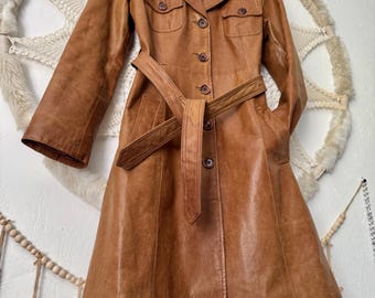 May include: A vintage brown leather trench coat with a belted waist, button closures, and two front pockets. The coat has a classic collar and long sleeves. The coat is hanging on a wooden hanger.