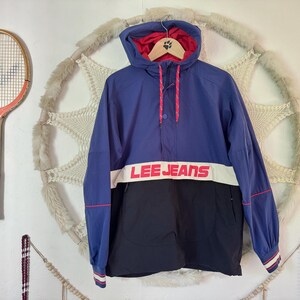 May include: A blue, black, and white Lee Jeans hooded jacket. The jacket has a red interior and trim, with the Lee Jeans logo across the chest. A vintage tennis racket hangs on the wall to the left.