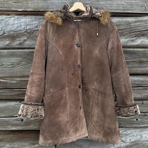 May include: A brown suede coat with a fur-lined hood and cuffs. The coat has a button-up front, two front pockets, and a tailored fit. The coat is hanging on a wooden hanger against a wooden background.