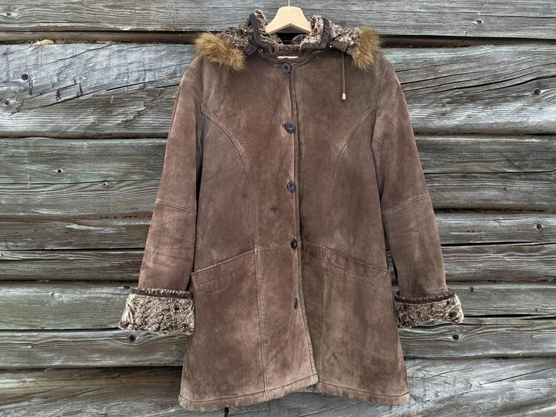 May include: A brown suede coat with a fur-lined hood and cuffs. The coat has a button-up front, two front pockets, and a tailored fit. The coat is hanging on a wooden hanger against a wooden background.