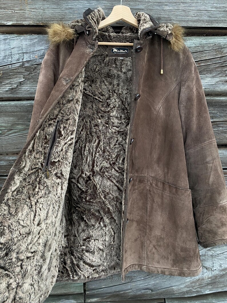 May include: A brown suede coat with a faux fur lining and hood. The coat has a button-up front and a label that reads "Maither". The hood is trimmed with faux fur.