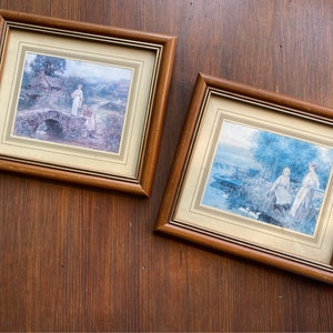 May include: Two framed pictures with brown wooden frames. The picture on the left shows a woman and a girl walking across a bridge in a rural setting. The picture on the right shows two women standing in a field with a lake in the background.