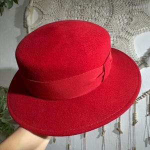 Op de afbeelding: Een felrode hoed met een brede rand en een cilindrische kroon. De hoed heeft een bijpassende rode lintband rond de kroon. De hoed is gemaakt van een viltachtig materiaal en is een stijlvol accessoire.