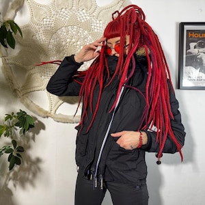 May include: A black jacket with a white zip and a black hood. The person in the image has red dreadlocks and is wearing red sunglasses. A framed poster with the word "Houston" is visible in the background.
