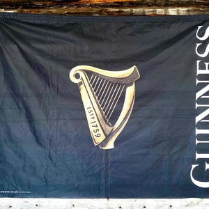以下が含まれることがあります： 黒い布の旗に、金のハープと「GUINNESS ESTD 1759」の文字が印刷されています。