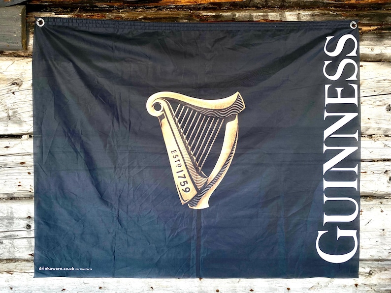 以下が含まれることがあります： 黒い布の旗に、金のハープと「GUINNESS ESTD 1759」の文字が印刷されています。